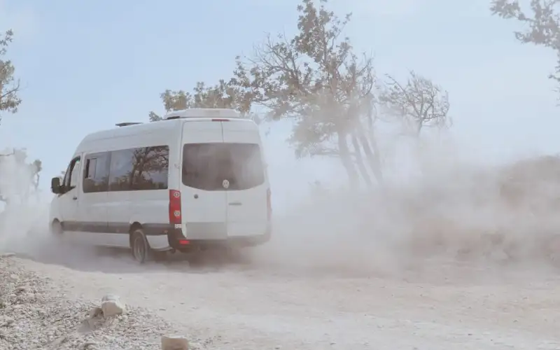 White passenger van driving down a dusty gravel road past dry trees