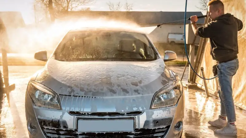 Man pressure washing a foam-covered car in golden light
