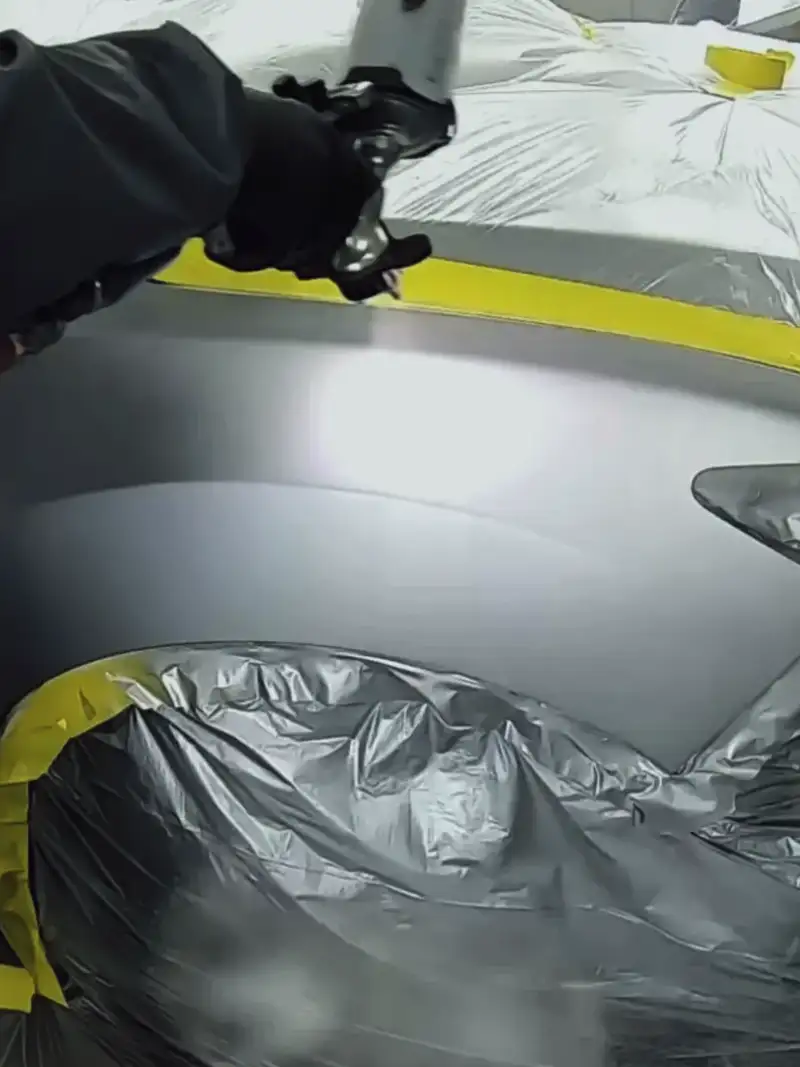 Painter in grey overalls spraying the masked front fender of a silver car in a paint booth